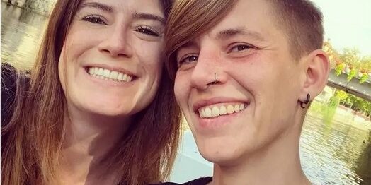 Eine dunkelhaarige Frau und eine blonde Frau mit kurzen Haaren schauen mit breitem Lächeln in Kamera. Im Hintergrund ist Neckar in Tübingen zu sehen.