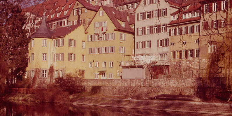 Blick auf eine Häuserzeile. Die Häuser spiegeln sich im Neckar. Das Haus am linken Bildrand hat einen kleinen runden Turm mit einem spitzen Dach.