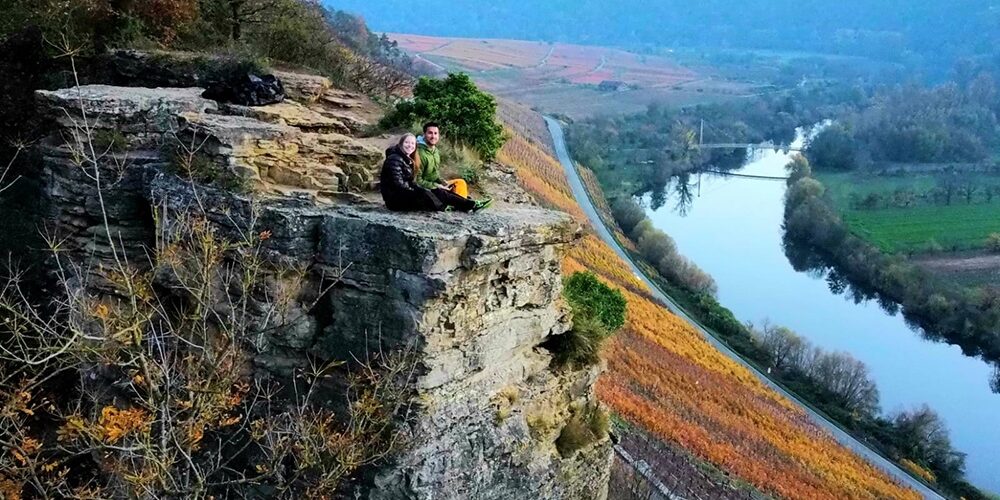 Zu sehen sind eine junge blonde Frau und ein dunkelhaariger Mann, welche links im Bild auf einem Felsen sitzen. Rechts sind der Neckar und die Weinberge zu sehen.