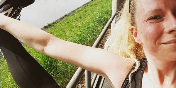 Eine blonde Frau hält ihr rechtes Bein mit der rechten Hand hoch und guckt in die Kamera. Im Hintergrund ist der Neckar neben dem Laufweg zu sehen.