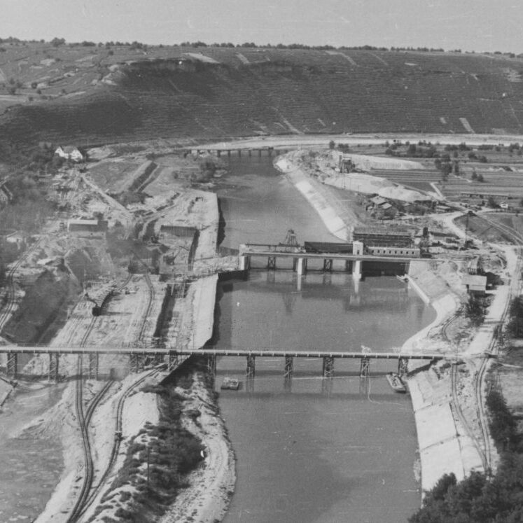 Blick auf eine große Baustelle mitten im Fluss. An beiden Ufern sind Eisenbahnschienen zu sehen.