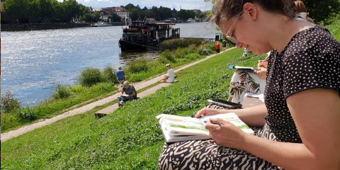 Eine dunkelhaarige Frau sitzt auf der Neckarwiese und malt in einem kleinen Heft - Im Hintergrund fließt der Neckar.