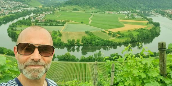 Ein Mann mit Sonnenbrille schaut in Kamera. Im Hintergrund ist die Neckarschleife in Mundelsheim zu sehen.
