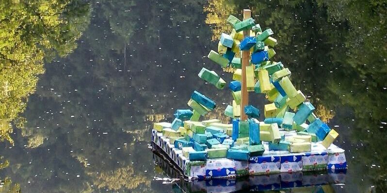 Auf dem Bild ist der Neckar zu sehen, wie er auf beiden Seiten von Gebüsch umrahmt wird. Entlang des Flusses fließen drei bunte plastische Kunstwerke auf Flößen.