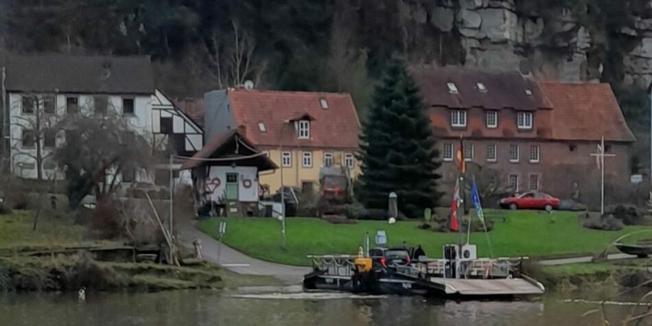 Eine kleine Fähre legt am Ufer an. Auf der Fähre ist ein schwarzes Auto. Im Hintergrund stehen Wohnhäuser.