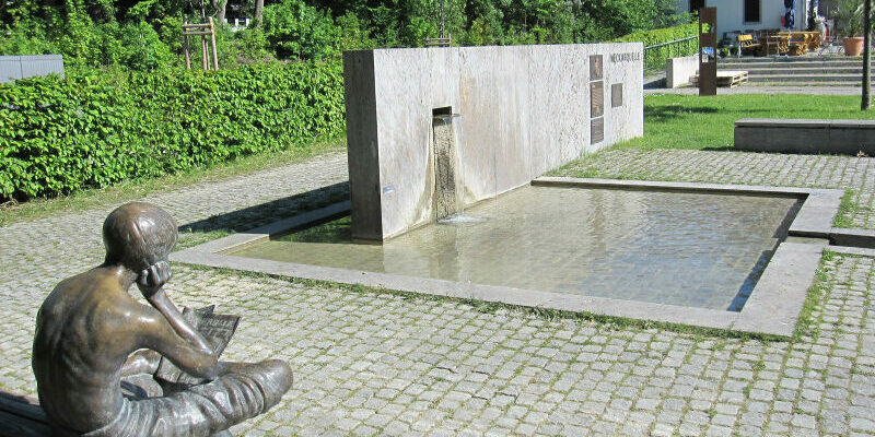 Neckarquelle im Stadtpark Möglingshöhe Skulptur eines Zeitung lesenden Jungen sitzt vor einem Wasserbecken. In das Becken fließt das Wasser der Neckarquelle aus einem rechteckigen Stein.