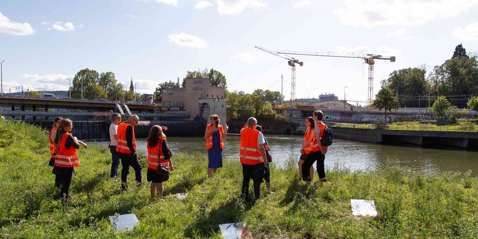 Eine Gruppe Menschen steht auf einer Insel am Neckar und hört einem Vortrag zu.
